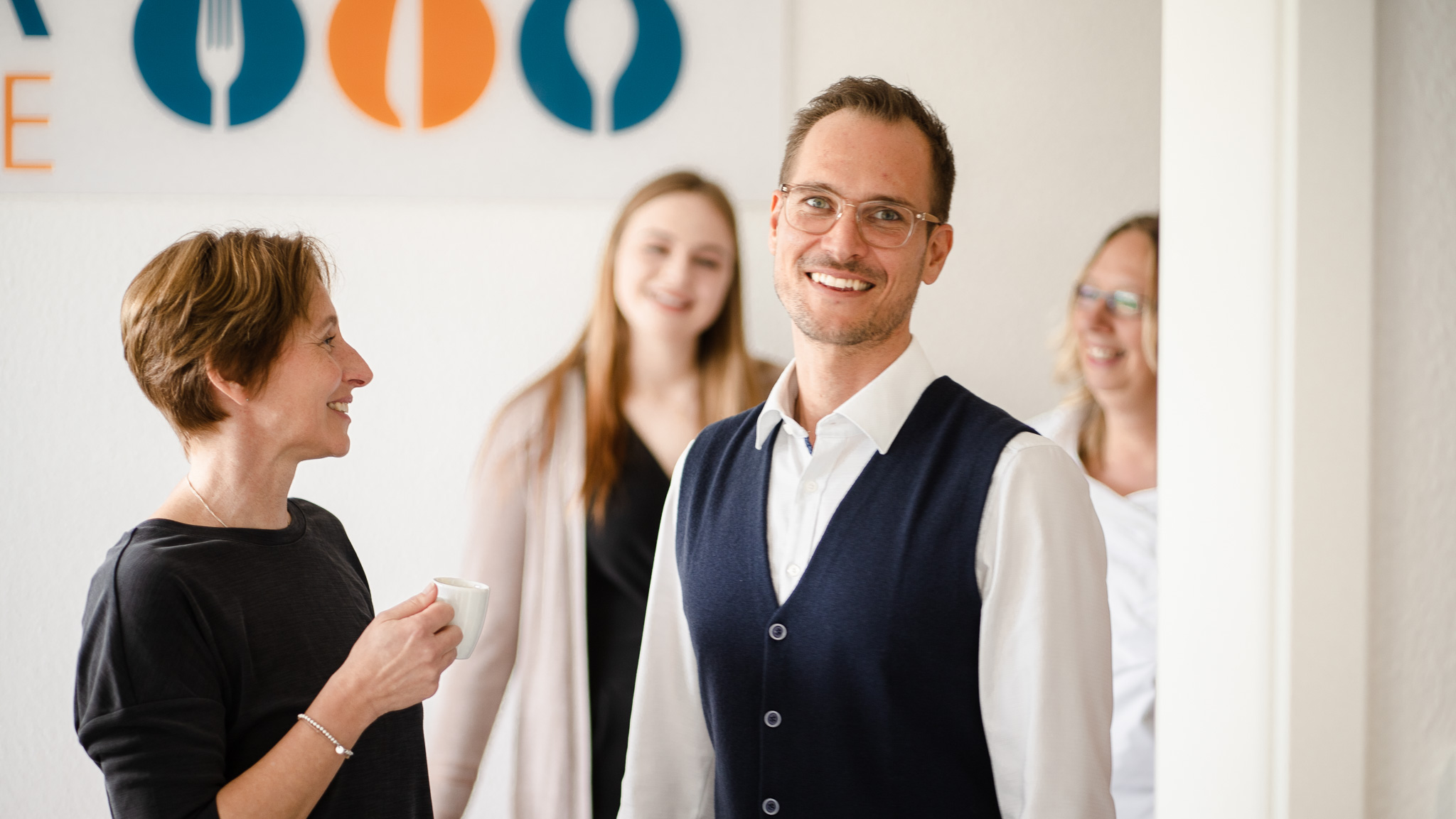 Mann steht im Vordergrund, lacht in die Kamera. Frauen verschwommen im Hintergrund.